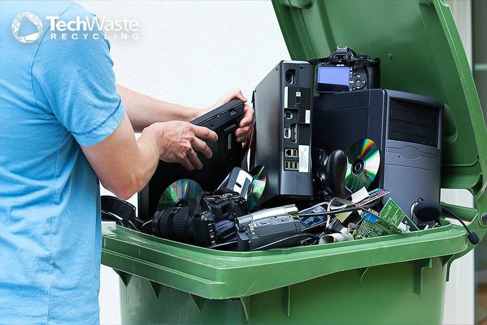 Person placing electronic devices such as computers, cameras, and cables in a green recycling bin, with the TechWaste Recycling logo in the corner.