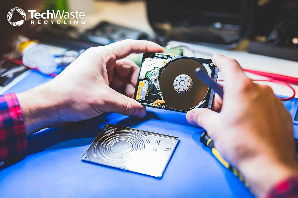 A person uses a tool to examine an opened hard drive on a workbench, with the TechWaste Recycling logo in the upper left corner.