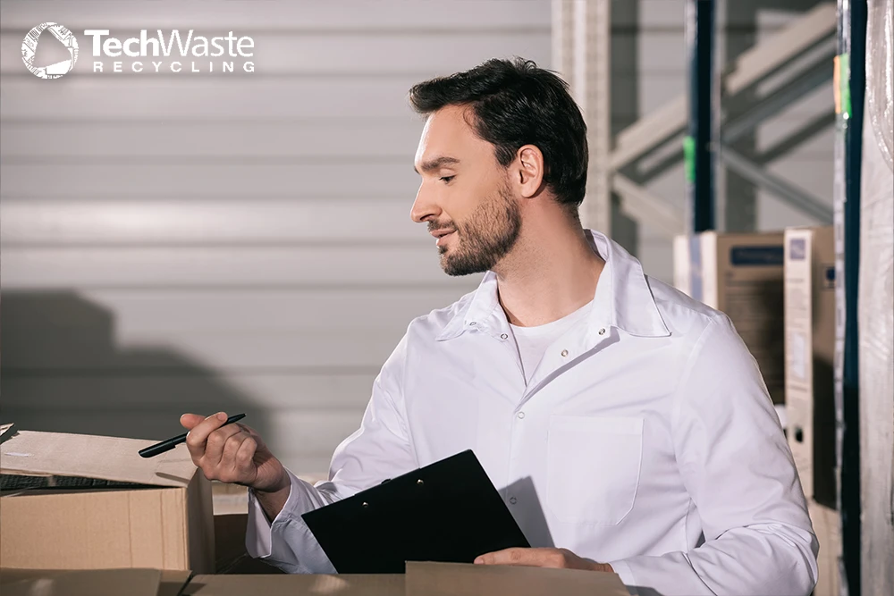 A man in a white lab coat holds a clipboard and pen, standing among cardboard boxes in a warehouse with "TechWaste Recycling" logo in the top left corner.