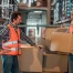 Two men in safety vests and plaid shirts stand in a warehouse; one holds a cardboard box, the other rests his hand on stacked boxes. "TechWaste Recycling" logo is visible.
