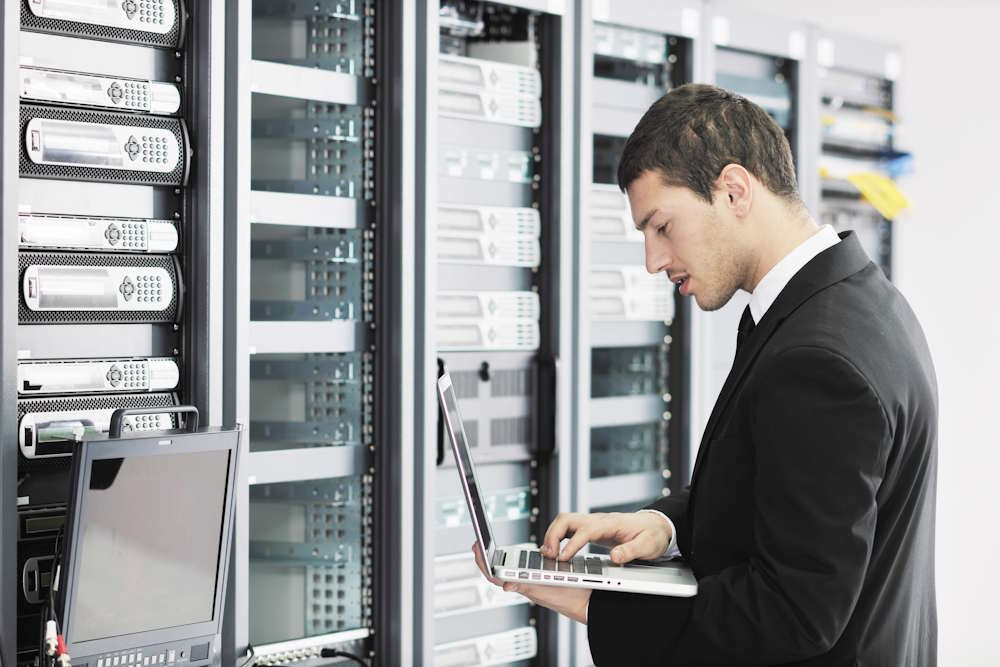 A man in a suit uses a laptop while standing in front of server racks in a data center.