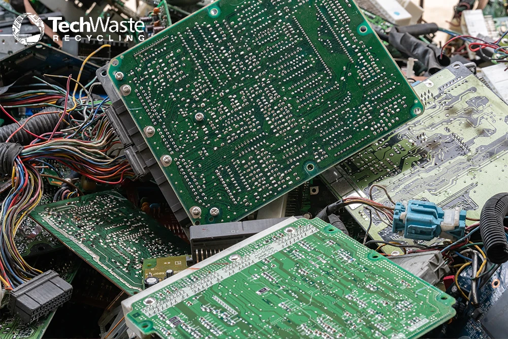 A pile of discarded circuit boards and electronic components, with wires tangled around them. TechWaste Recycling logo is visible in the top left corner.
