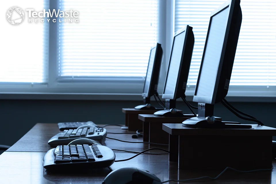 Three desktop computers with monitors, keyboards, and mice are arranged in a row on a desk in front of windows with blinds. The "TechWaste Recycling" logo is visible in the corner.