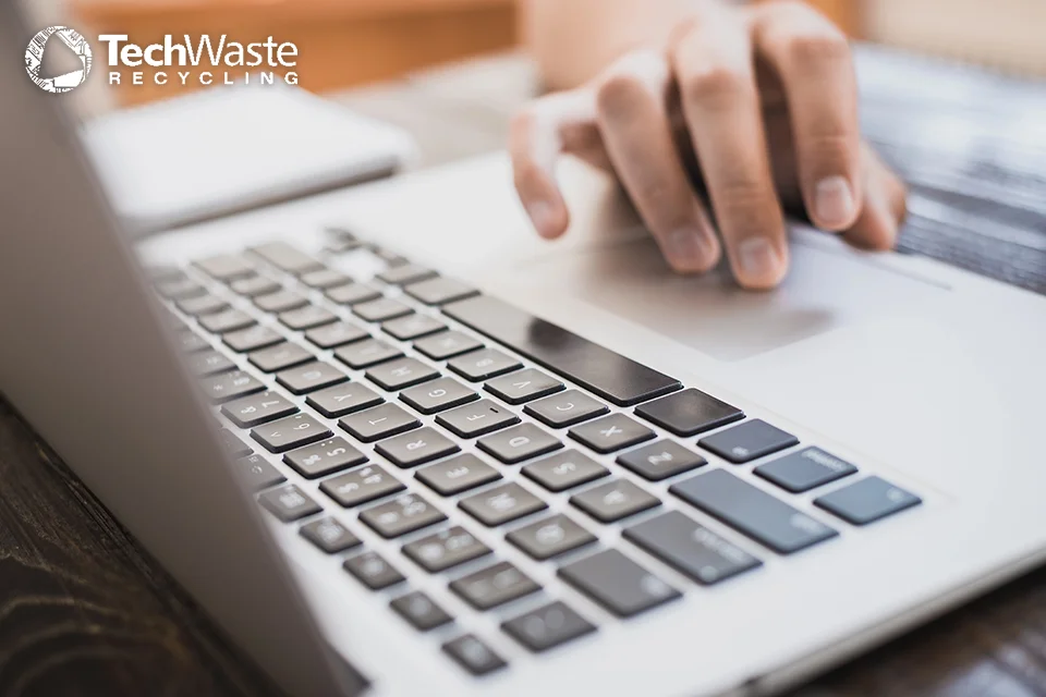 A hand using the trackpad of an open laptop, with the TechWaste Recycling logo visible in the upper left corner.