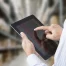 A person uses a tablet in a warehouse, tapping the screen with one finger while holding the device in their other hand. Shelves and boxes are blurred in the background.
