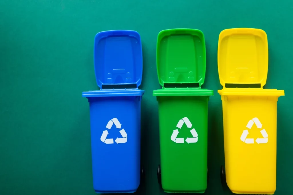Three recycling bins in blue, green, and yellow with lids open, placed side by side on a green background, each displaying the recycling symbol on the front.
