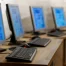Four desktop computers with keyboards and monitors are arranged in a row on a wooden desk, all screens displaying blurred content.