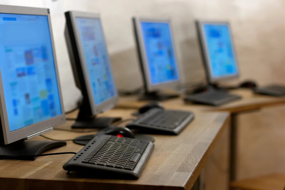 Four desktop computers with keyboards and monitors are arranged in a row on a wooden desk, all screens displaying blurred content.