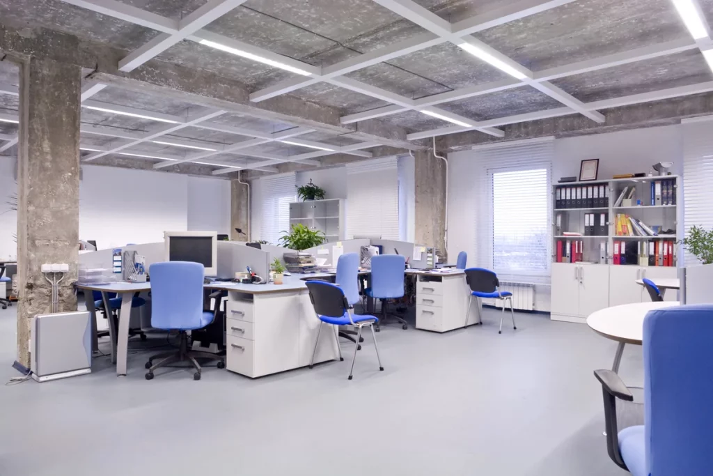 Modern open-plan office with several workstations, blue chairs, computers, and shelves with files and plants. Large windows let in natural light. Room appears clean and organized.