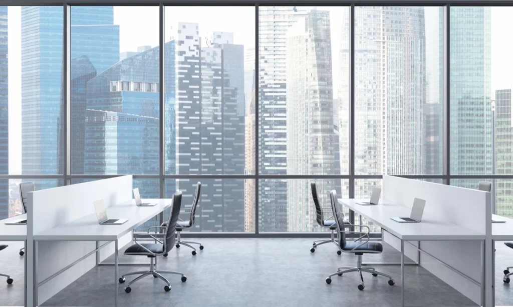 Modern office space with desks, chairs, and laptops, featuring large windows overlooking a cityscape of tall skyscrapers.
