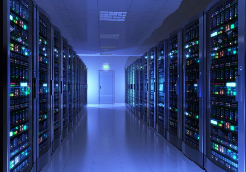 A brightly lit data center room with rows of server racks on both sides, visible cables, and a closed door at the far end.