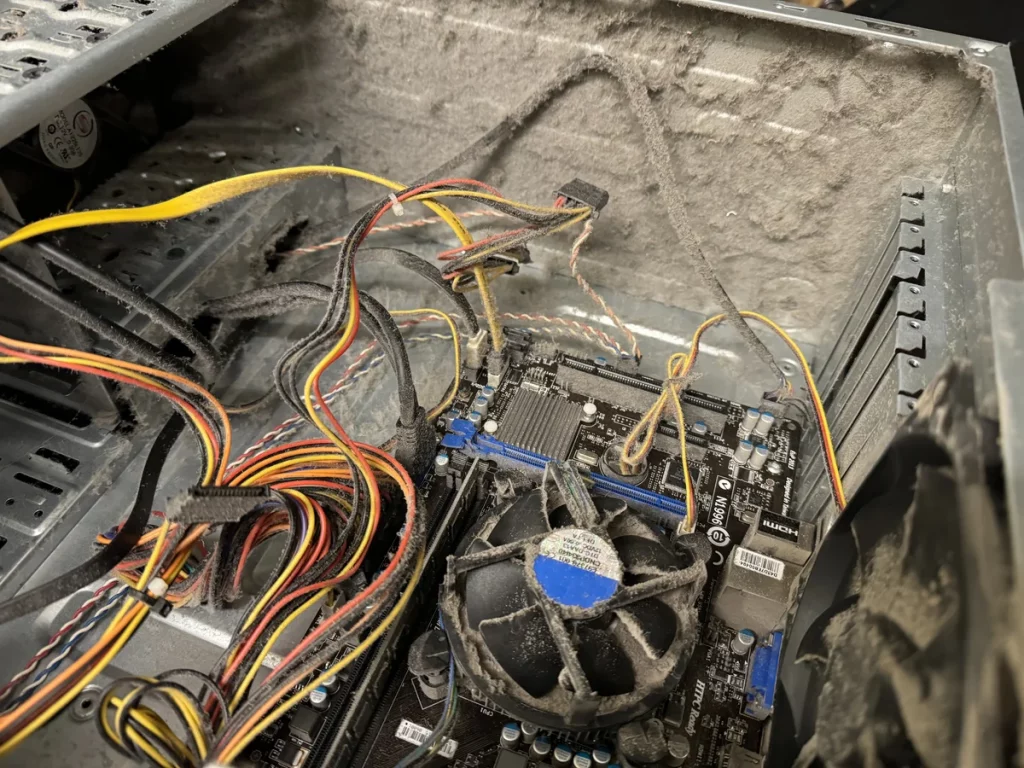The inside of a dusty desktop computer showing exposed cables, a motherboard, and a fan covered in dust buildup.