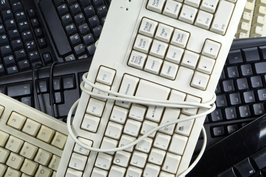 A pile of old computer keyboards, some black and some white, with one white keyboard wrapped in its cord lying on top.