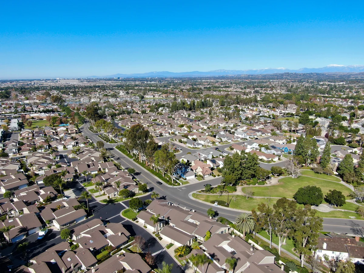 Aerial view of a suburban neighborhood with rows of houses, streets, and a green park area under a clear blue sky.