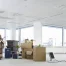 A person unpacking boxes in a bright, empty office with large windows, scattered boxes, a phone, and a laptop on the floor.