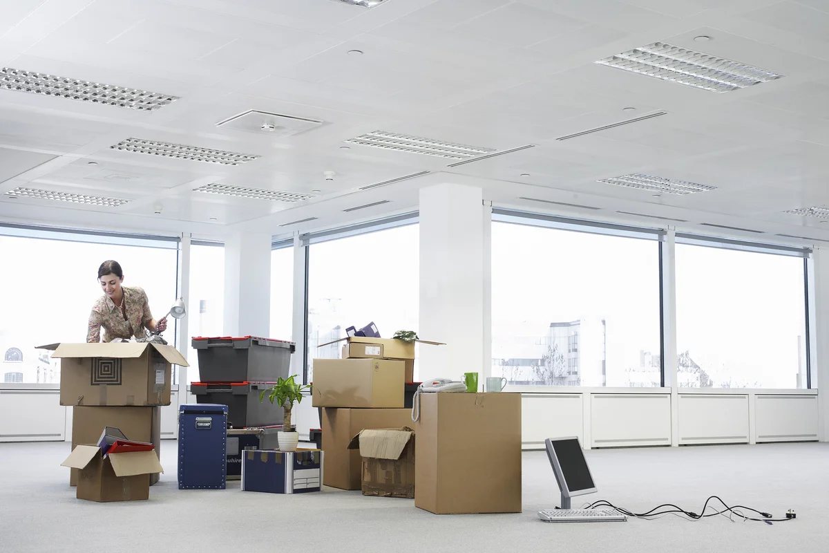 A person unpacking boxes in a bright, empty office with large windows, scattered boxes, a phone, and a laptop on the floor.