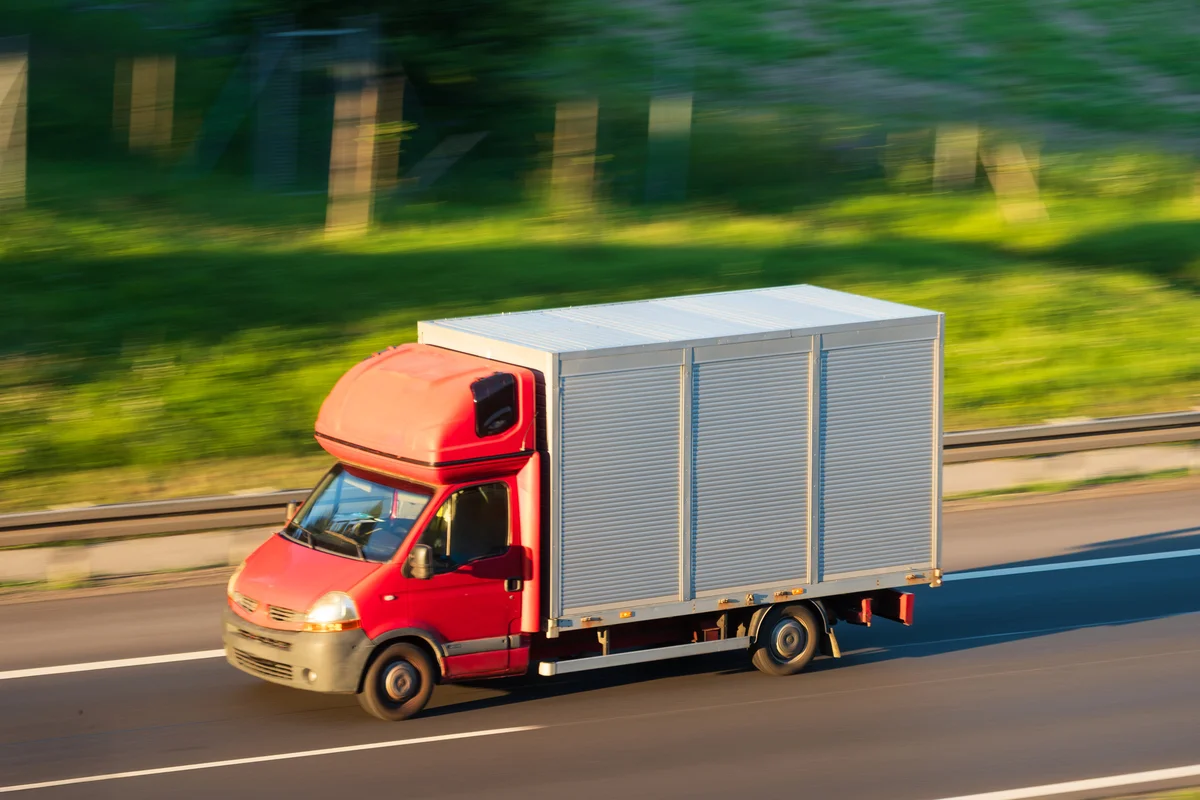 Red box truck driving on a highway in daylight, with a blurred green background indicating motion.